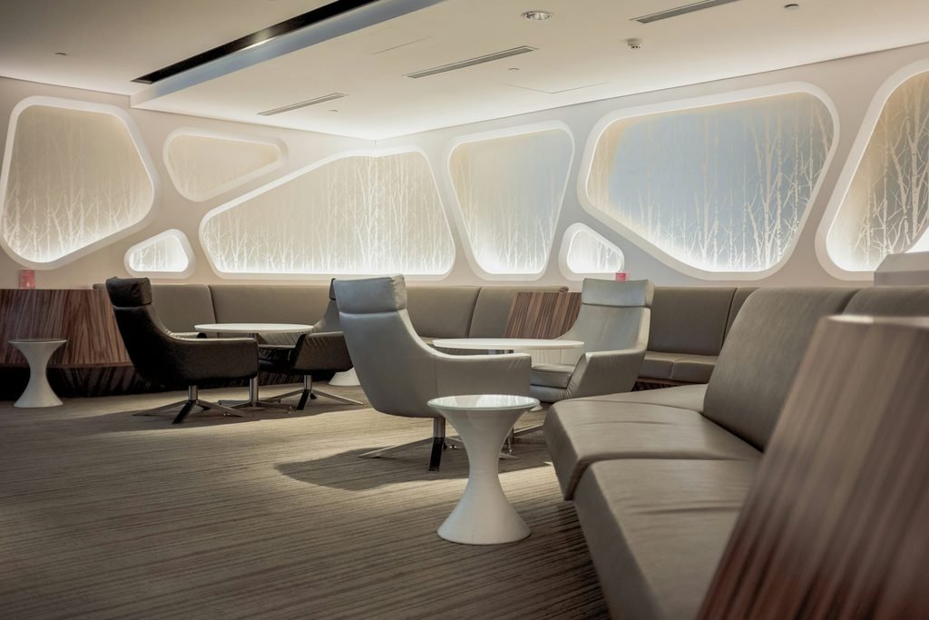 Lounge de aeroporto com poltronas, sofás e mesas em tons claros.